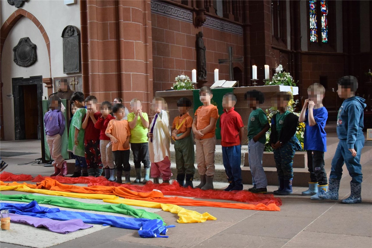 Eine Kindergruppe steht in einer Kirche vor dem Altar, bunt gekleidet. Vor ihnen liegt ein bunter Regenbogen aus farbigen Tüchern.