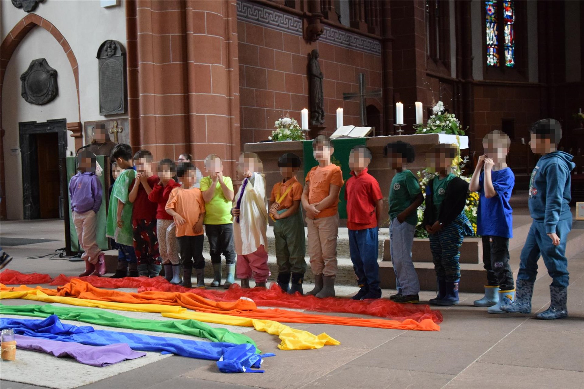 Eine Kindergruppe steht in einer Kirche vor dem Altar, bunt gekleidet. Vor ihnen liegt ein bunter Regenbogen aus farbigen Tüchern.