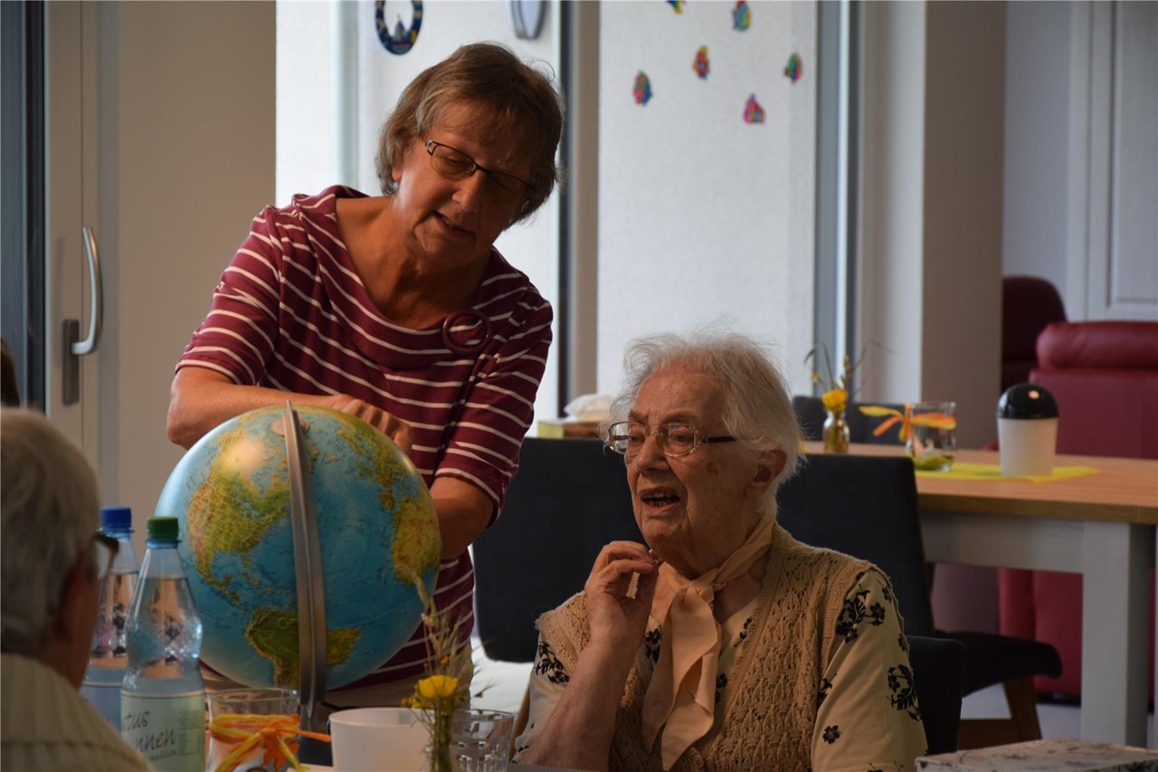 Eine Mitarbeiterin der Tagespflege im Gespräch mit einer Seniorin, die an einem Tisch sitzt. Beide schauen sich eine Weltkugel an und zeigen auf Länder.