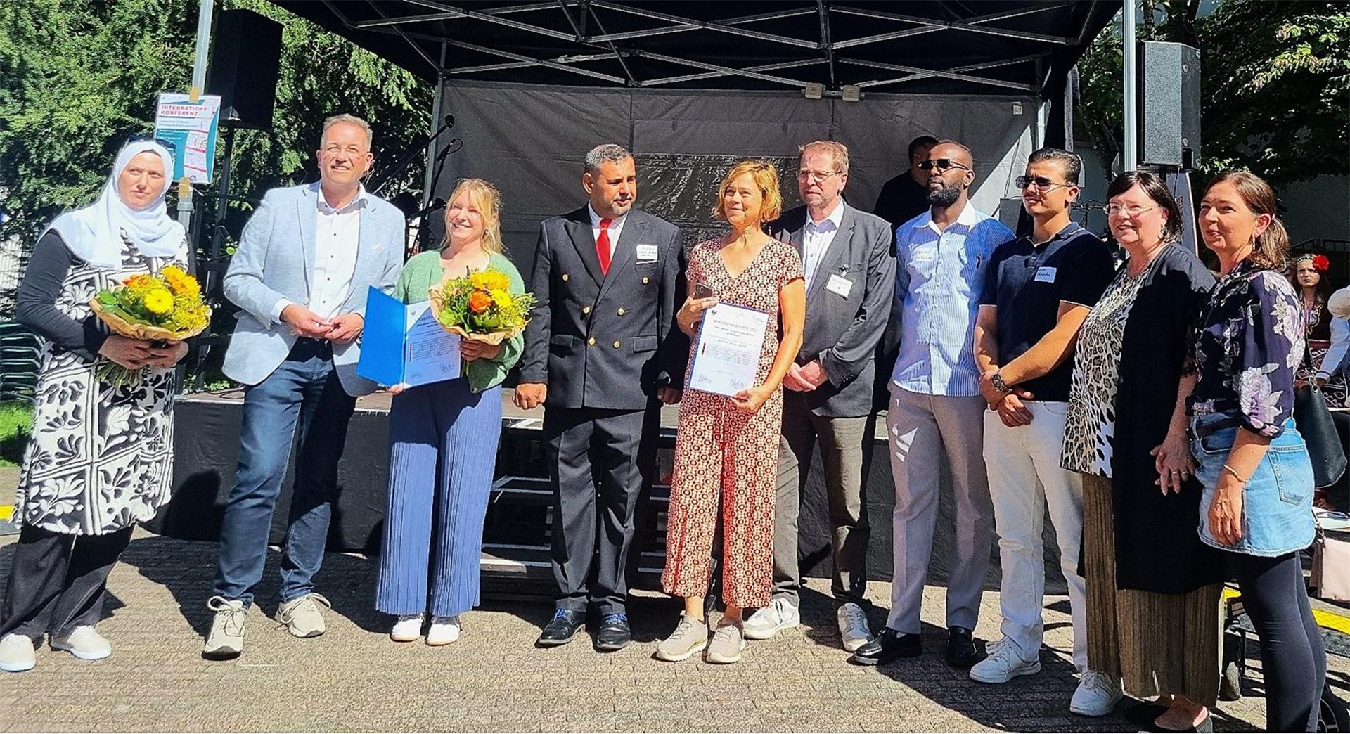 Mehrere Männer und Frauen stehen im Rahmen einer Preisverleihung für ein Gruppenbild zusammen. Einige Personen halten Blumensträuße und Urkunden in der Hand.