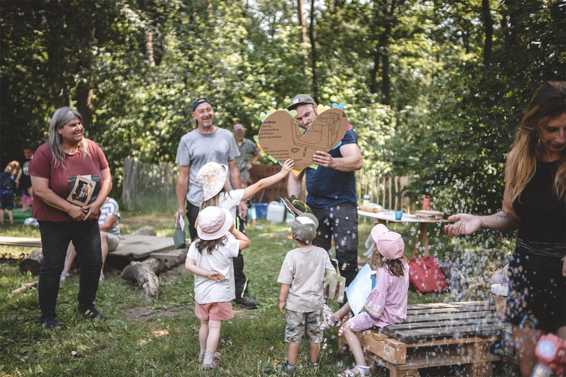 Eltern und Kinder stehen inmitten Seifenblasen zusammen im Wald. Ein Mann hält ein Herz aus Passe in die Höhe.