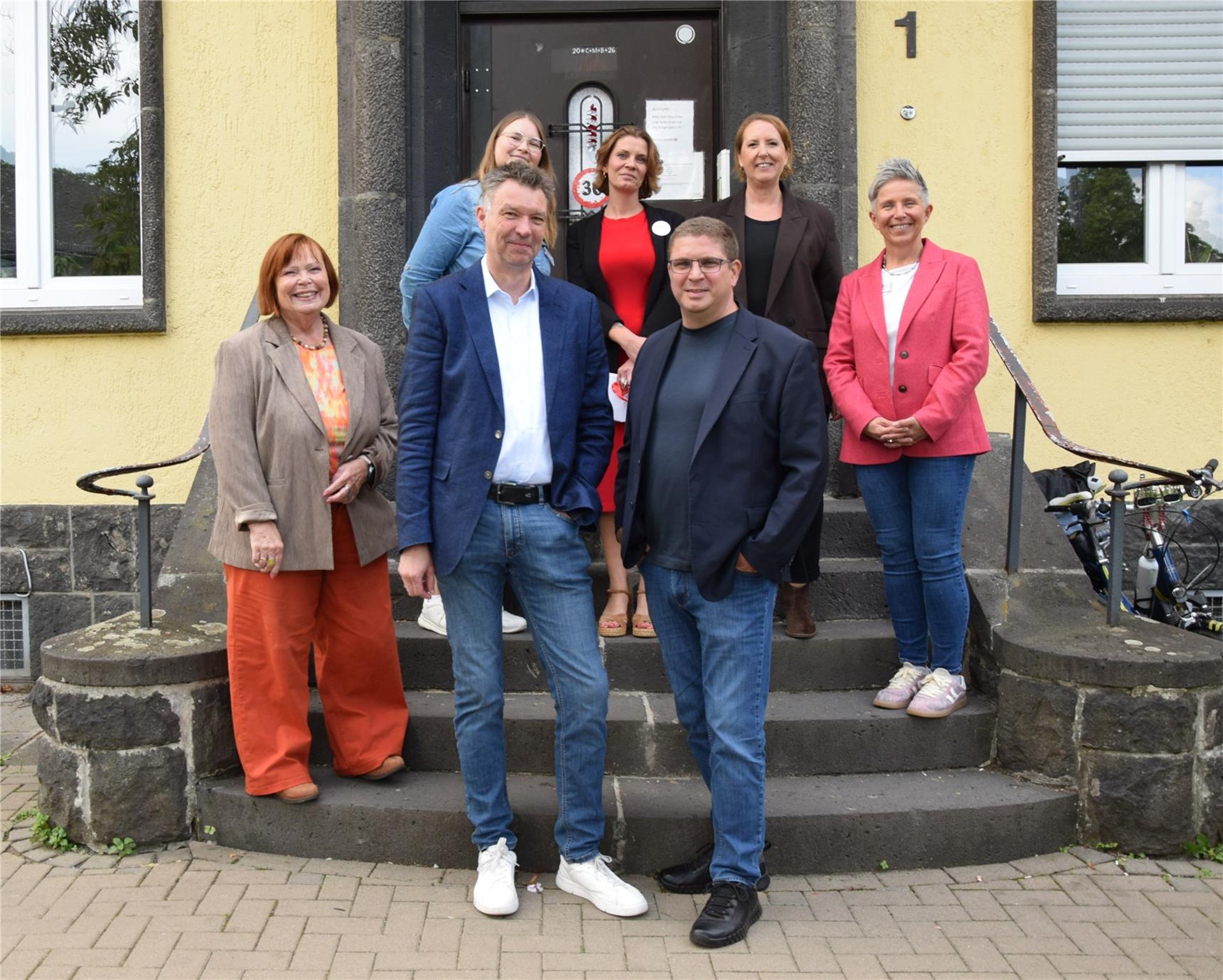 Mehrere Personen stehen auf einer Treppe vor einem Haus zu einem Gruppenbild zusammen.